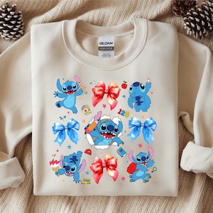 Op de afbeelding: Een beige sweater met ronde hals en een grafische print van het blauwe personage Stitch in verschillende houdingen, waaronder met een potlood, een kwast en een rugzak. Het ontwerp bevat rode en blauwe strikken, kleine sterren en schooliconen.