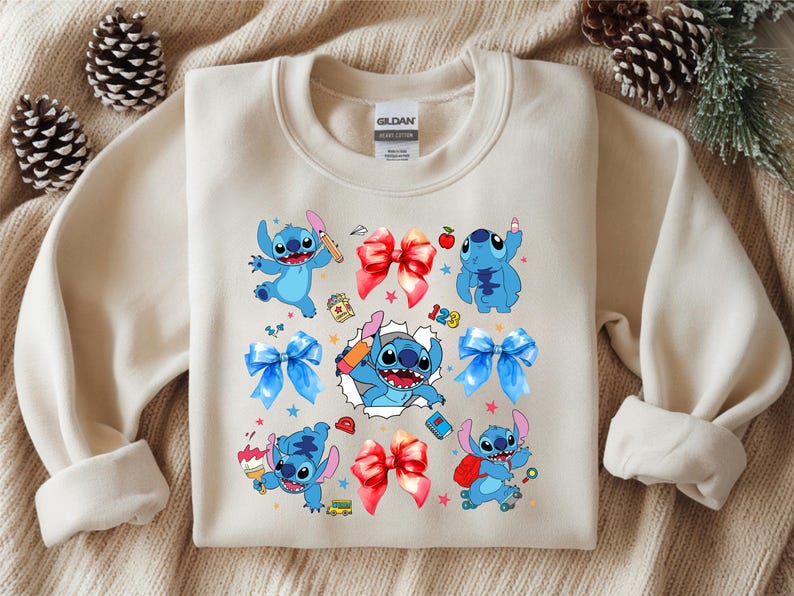 Op de afbeelding: Een beige sweater met ronde hals en een grafische print van het blauwe personage Stitch in verschillende houdingen, waaronder met een potlood, een kwast en een rugzak. Het ontwerp bevat rode en blauwe strikken, kleine sterren en schooliconen.