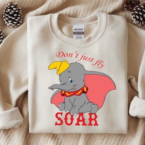May include: Cream-colored sweatshirt featuring a graphic of Dumbo the elephant with a yellow hat, red collar, and pink ears. The text "Don't just fly SOAR" is printed in red. A cozy, casual top.