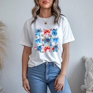Op de afbeelding: Een wit t-shirt met korte mouwen voorzien van een herhalende grafische print van het blauwe personage Stitch uit Lilo en Stitch, afgewisseld met rode en blauwe strikken, kleine rode appels en diverse kleine kleurrijke iconen op de borst.