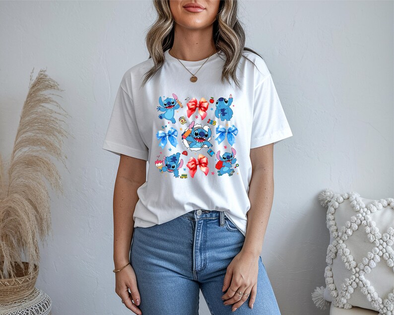 Op de afbeelding: Een wit t-shirt met korte mouwen voorzien van een herhalende grafische print van het blauwe personage Stitch uit Lilo en Stitch, afgewisseld met rode en blauwe strikken, kleine rode appels en diverse kleine kleurrijke iconen op de borst.