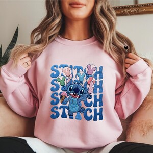 May include: A light pink sweatshirt featuring a blue cartoon character holding ice cream and balloons. The word "STITCH" is printed in blue, stacked vertically. The design is centered on the front of the sweatshirt.