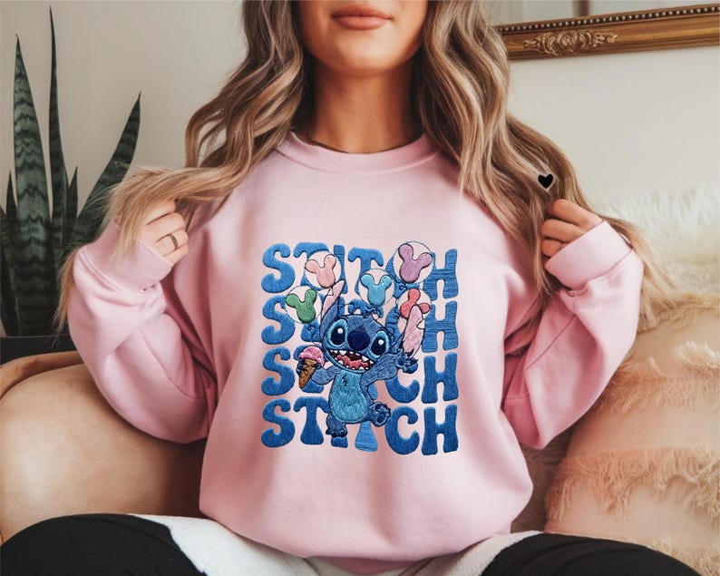 May include: A light pink sweatshirt featuring a blue cartoon character holding ice cream and balloons. The word "STITCH" is printed in blue, stacked vertically. The design is centered on the front of the sweatshirt.