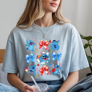 Op de afbeelding: Een lichtblauw t-shirt met een grafische print van het blauwe personage Stitch in verschillende houdingen, afgewisseld met rode en blauwe strikken, kleine schoolgerelateerde iconen en een centraal beeld van Stitch die door een witte uitsnede breekt.