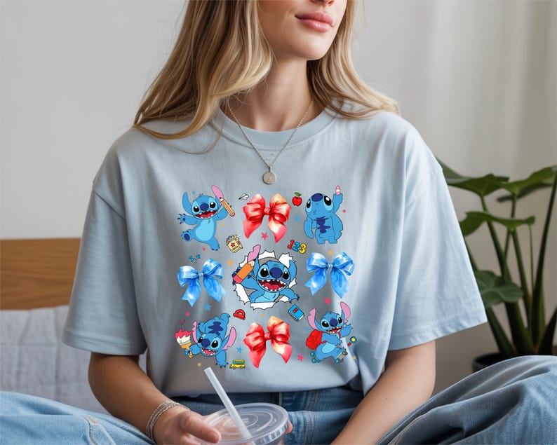 Op de afbeelding: Een lichtblauw t-shirt met een grafische print van het blauwe personage Stitch in verschillende houdingen, afgewisseld met rode en blauwe strikken, kleine schoolgerelateerde iconen en een centraal beeld van Stitch die door een witte uitsnede breekt.