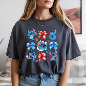 Op de afbeelding: Een antracietgrijs T-shirt met korte mouwen en een grafische print van het blauwe personage Stitch in verschillende houdingen, afgewisseld met rode en blauwe strikken, kleine kleurrijke iconen en een centrale afbeelding van Stitch die uit een witte scheur komt.