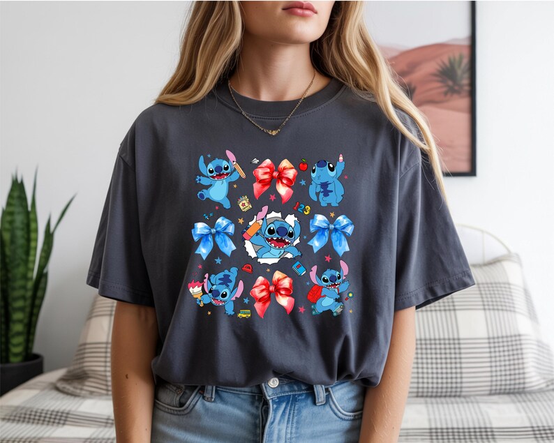 Op de afbeelding: Een antracietgrijs T-shirt met korte mouwen en een grafische print van het blauwe personage Stitch in verschillende houdingen, afgewisseld met rode en blauwe strikken, kleine kleurrijke iconen en een centrale afbeelding van Stitch die uit een witte scheur komt.