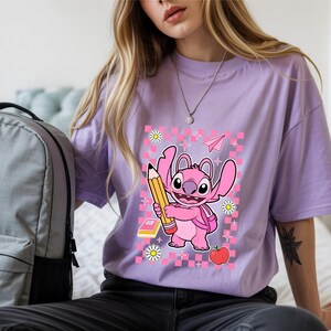 Może przedstawiać: Jasnofioletowy t-shirt z grafiką przedstawiającą r&oacute;żowego kosmitę trzymającego duży ż&oacute;łty oł&oacute;wek. Wz&oacute;r zawiera r&oacute;żowe tło w kratkę, czerwone jabłko, małą książkę, białe stokrotki oraz ikonę r&oacute;żowego papierowego samolotu.