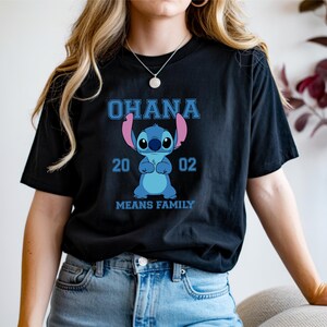 May include: Black t-shirt featuring a blue cartoon character with the text "OHANA 2002 MEANS FAMILY". The character is blue with large eyes and pink ears. The text is in a blue, blocky font.