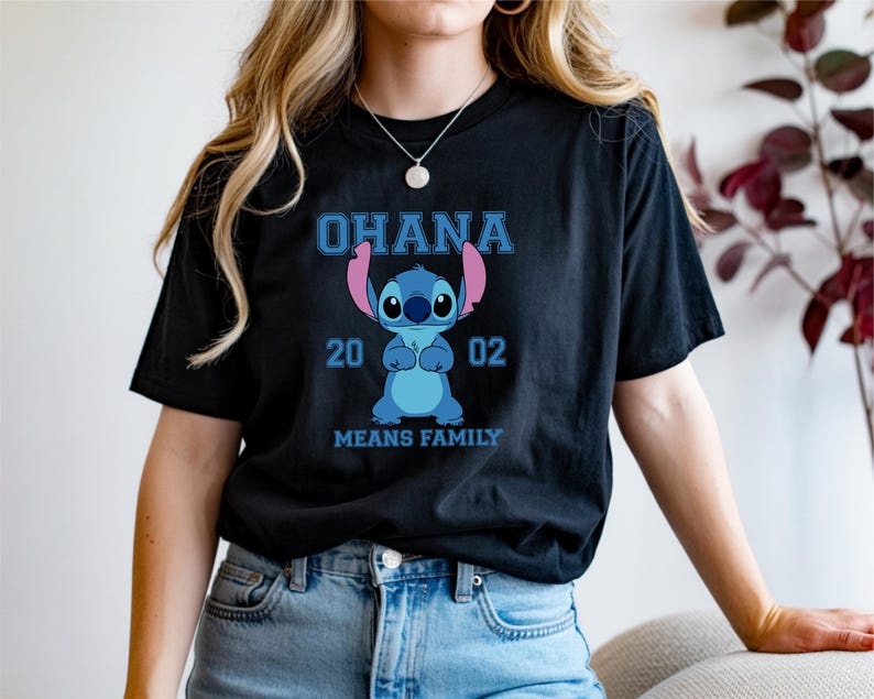 May include: Black t-shirt featuring a blue cartoon character with the text "OHANA 2002 MEANS FAMILY". The character is blue with large eyes and pink ears. The text is in a blue, blocky font.
