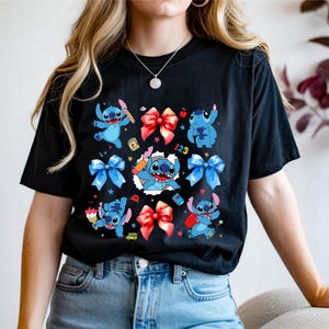 Op de afbeelding: Een zwart t-shirt met korte mouwen voorzien van een grafische print van het blauwe personage Stitch in verschillende houdingen, afgewisseld met rode en blauwe strikken, kleine sterretjes en schoolgerelateerde items zoals een potlood, een boek en de cijfers 123.