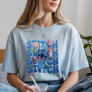 May include: Light blue t-shirt featuring the character Stitch from Lilo & Stitch. The design includes Stitch holding ice cream and balloons, with the name "STITCH" in a stacked, bubble-style font. The shirt is a casual, short-sleeved style.
