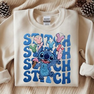 May include: Cream-colored sweatshirt featuring a blue embroidered Stitch graphic holding an ice cream cone and balloons. The word "STITCH" is embroidered in blue vertically down the left side of the graphic. The sweatshirt has long sleeves.
