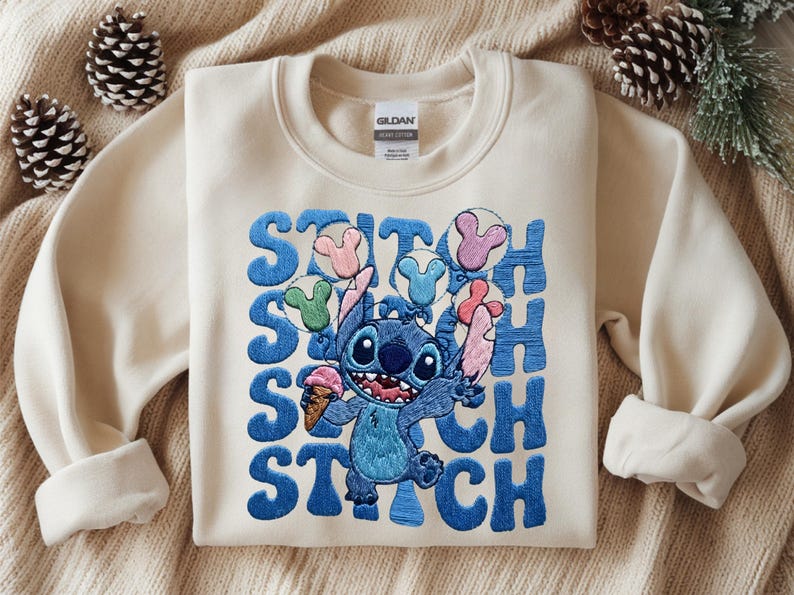 May include: Cream-colored sweatshirt featuring a blue embroidered Stitch graphic holding an ice cream cone and balloons. The word "STITCH" is embroidered in blue vertically down the left side of the graphic. The sweatshirt has long sleeves.
