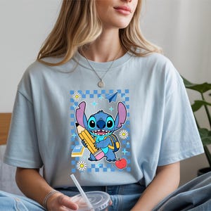 Może przedstawiać: Jasnoniebieski t-shirt z grafiką przedstawiającą niebieską postać Stitcha trzymającego duży ż&oacute;łty oł&oacute;wek. Wz&oacute;r zawiera niebieskie tło w kratkę, czerwone jabłko, mały stos książek, papierowy samolot oraz białe kwiaty stokrotki.