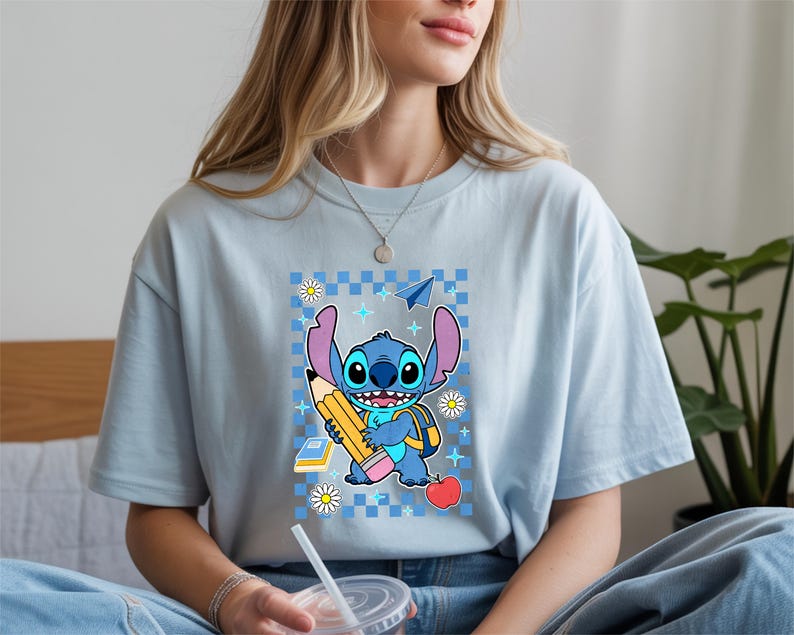 Może przedstawiać: Jasnoniebieski t-shirt z grafiką przedstawiającą niebieską postać Stitcha trzymającego duży ż&oacute;łty oł&oacute;wek. Wz&oacute;r zawiera niebieskie tło w kratkę, czerwone jabłko, mały stos książek, papierowy samolot oraz białe kwiaty stokrotki.