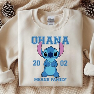 May include: Cream-colored sweatshirt featuring a blue cartoon character with large ears and the text "OHANA" above it. Below the character are the numbers "20 02" and the phrase "MEANS FAMILY".