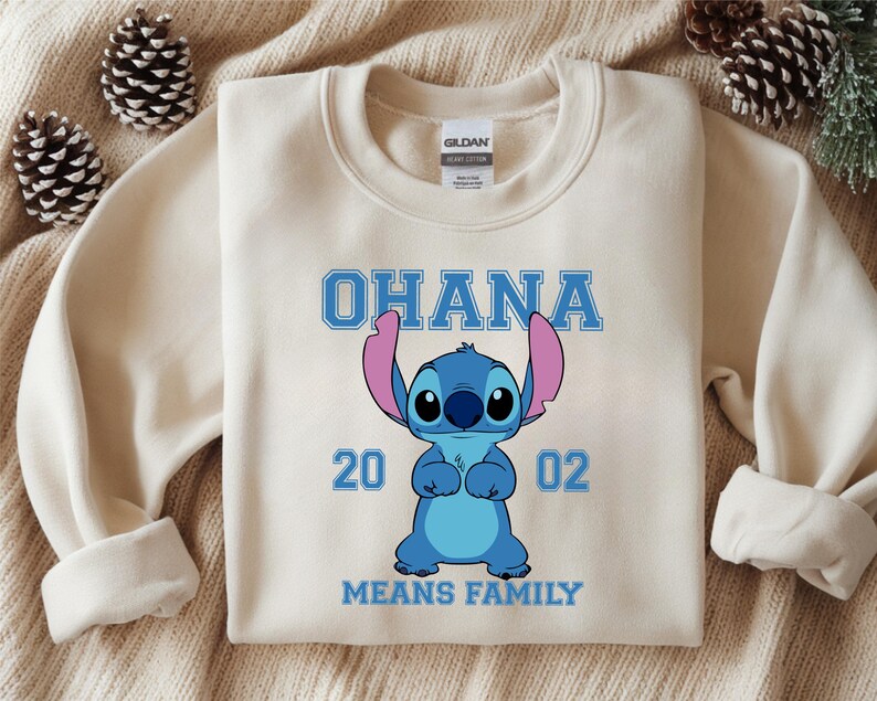 May include: Cream-colored sweatshirt featuring a blue cartoon character with large ears and the text "OHANA" above it. Below the character are the numbers "20 02" and the phrase "MEANS FAMILY".