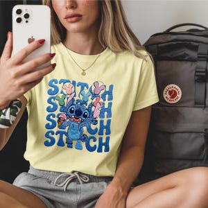 May include: A light yellow t-shirt featuring the character Stitch from Lilo & Stitch. The blue Stitch graphic is holding ice cream and is surrounded by the word "Stitch" in a repeating pattern. A black backpack is in the background.