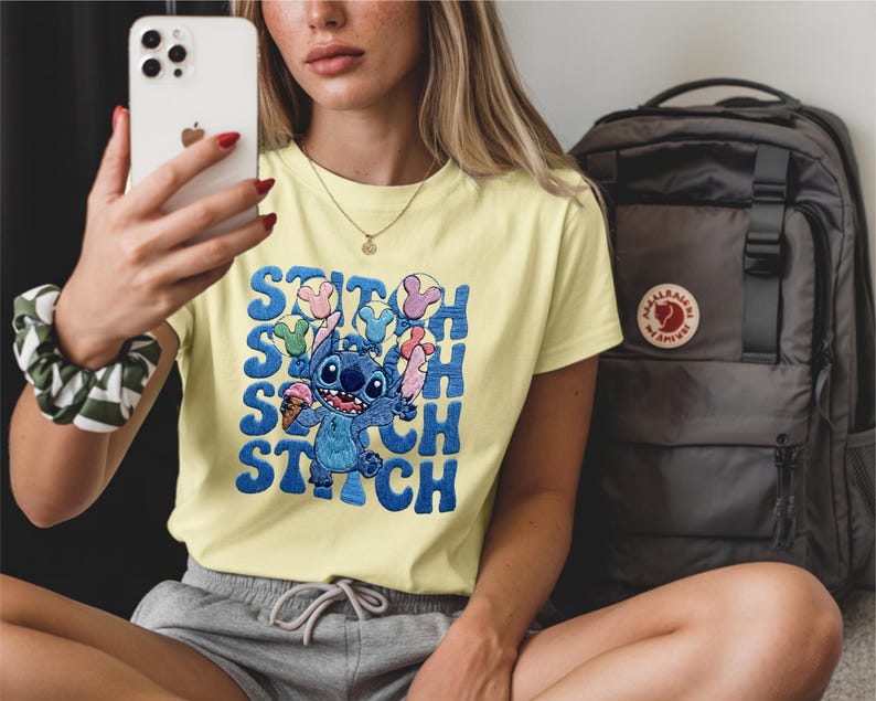 May include: A light yellow t-shirt featuring the character Stitch from Lilo & Stitch. The blue Stitch graphic is holding ice cream and is surrounded by the word "Stitch" in a repeating pattern. A black backpack is in the background.
