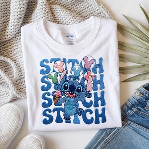 May include: White t-shirt featuring a blue Stitch graphic holding ice cream and balloons. The word "STITCH" is embroidered in blue. The shirt is paired with blue jeans and white sneakers.