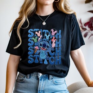 May include: Black t-shirt featuring a blue cartoon character holding an ice cream cone, with the word "STITCH" in blue block letters. The character is surrounded by colorful Mickey Mouse-shaped ice cream. The shirt is a casual, comfortable style.