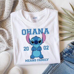 May include: White t-shirt featuring a blue cartoon character with large ears and the text "OHANA" above it. Below the character are the numbers "20 02" and the phrase "MEANS FAMILY" in blue.