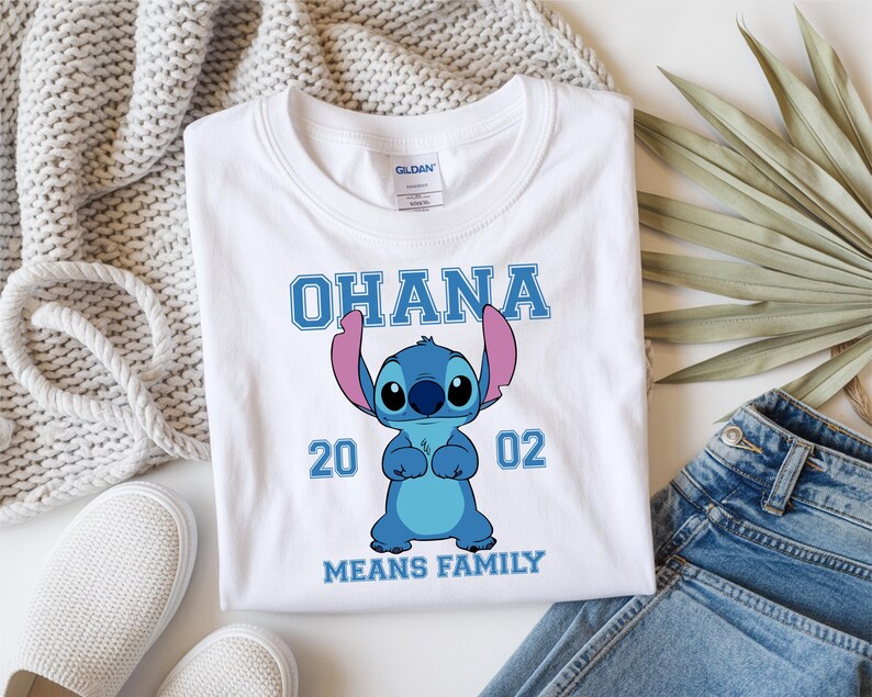 May include: White t-shirt featuring a blue cartoon character with large ears and the text "OHANA" above it. Below the character are the numbers "20 02" and the phrase "MEANS FAMILY" in blue.