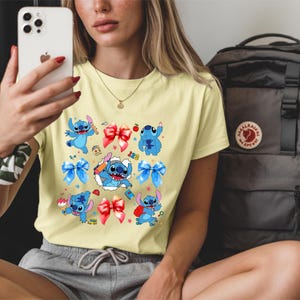 Op de afbeelding: Een lichtgeel t-shirt met korte mouwen en een grafische print van het blauwe personage Stitch in verschillende houdingen, afgewisseld met rode en blauwe strikken, kleine sterretjes, een appel, een potlood en de cijfers 1, 2 en 3 in kleurrijk lettertype.