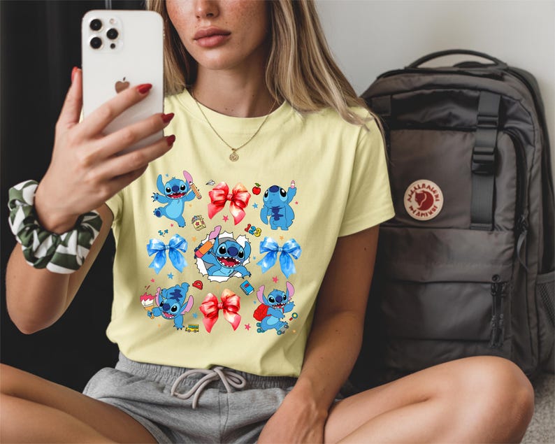 Op de afbeelding: Een lichtgeel t-shirt met korte mouwen en een grafische print van het blauwe personage Stitch in verschillende houdingen, afgewisseld met rode en blauwe strikken, kleine sterretjes, een appel, een potlood en de cijfers 1, 2 en 3 in kleurrijk lettertype.