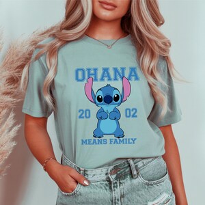 May include: A light blue t-shirt featuring a cartoon Stitch character with the word "OHANA" above and "MEANS FAMILY" below. The number "2002" is printed on the shirt. The shirt is made of a soft material.