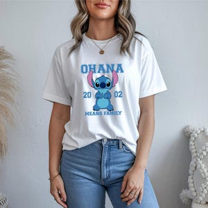 May include: White t-shirt featuring a blue cartoon character with the text "OHANA 2002 MEANS FAMILY". The graphic design is centered on the front of the shirt. The shirt is made of a soft, comfortable material.