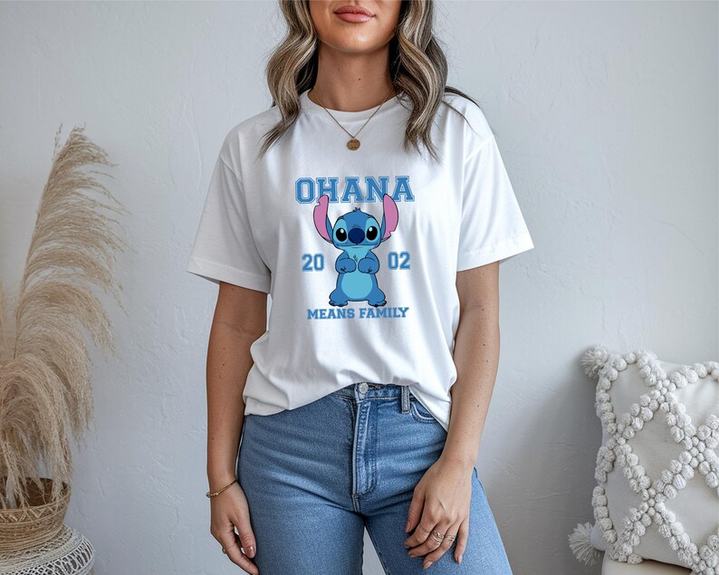 May include: White t-shirt featuring a blue cartoon character with the text "OHANA 2002 MEANS FAMILY". The graphic design is centered on the front of the shirt. The shirt is made of a soft, comfortable material.