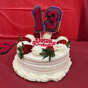 Op de afbeelding: Een ronde witte taart met witte frosting en verse aardbeien. De taart is bekroond met een rode en zilveren nummer 13 taarttopper en de woorden "Happy Birthday" in rode glazuur.