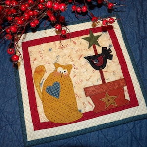 May include: A square, quilted wall hanging with a folk art design. It features a yellow cat with a blue heart, a black bird, and a star. The border is red and green, with a cream background.