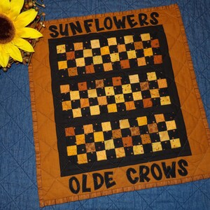 May include: A quilted wall hanging with the words "SUNFLOWERS" and "OLDE CROWS" in black lettering. The center features a black background with a checkerboard pattern in shades of yellow, orange, and brown. The border is a solid brown.
