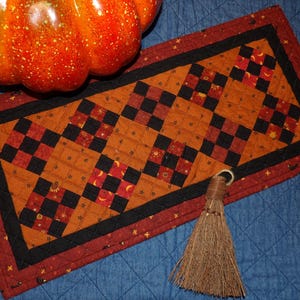 May include: A decorative autumn-themed table runner with a patchwork design in orange, black, and red. The runner features a central pattern of alternating squares and is bordered by a black and red trim. A small broom and a ceramic pumpkin are also in the image.