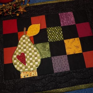May include: A quilted table runner with a patchwork design featuring black, red, green, yellow, and purple squares. A green and white checkered pear with a red heart is centered on the runner.