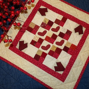 May include: A handmade quilted table topper with a heart and arrow design. The square topper features a cream-colored background with red hearts and arrows, and red and white polka dot borders. The quilt is approximately 18 inches square.