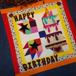 May include: A colorful, handmade birthday quilt with the words "HAPPY BIRTHDAY" in brown letters. The quilt features a cake with candles, star shapes, and geometric patterns in various colors. The border is red and orange.