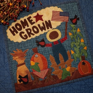 May include: A square fabric wall hanging featuring a blue checkered background with the text HOME GROWN in brown letters. The design includes a scarecrow, a pumpkin, corn stalks, a small American flag, and a black bird on a dark blue quilt.