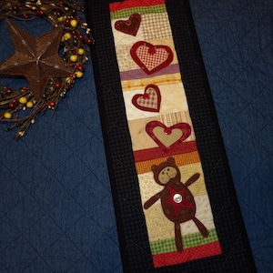 May include: A quilted table runner with a black border and a design of hearts and a teddy bear. The hearts are in shades of red and brown, and the teddy bear is brown with a red star on its chest.