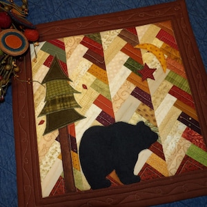 May include: A framed quilted wall hanging with a brown frame and a patchwork design. The design features a black bear silhouette, a green tree, and a yellow crescent moon with a red star.