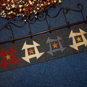 May include: A rectangular fabric wall hanging featuring a dark blue background with a small red floral pattern. It displays four appliqued star motifs in red, cream, blue plaid, and cream, each centered with a small orange star, suspended from a black metal rack.