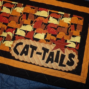 May include: A black quilted table runner with an orange and brown striped border. The center features a brown and tan applique with the text "CAT-TAILS" in black letters. The background is a pattern of orange, brown, and white cats.