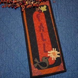 May include: A rectangular quilted wall hanging featuring a vertical orange panel with the word FALL appliqued in dark red letters. The design includes a black bird, a tan plaid star, and a green leaf accent on a black background with orange borders.