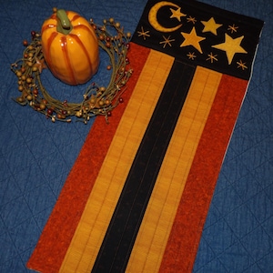May include: A decorative fall-themed table runner with a black top section featuring a crescent moon and stars. The runner has orange, yellow, and black vertical stripes. A ceramic pumpkin and a berry wreath are also in the image.