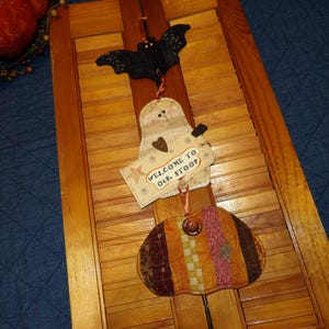 May include: A Halloween-themed hanging decoration featuring a black bat, a ghost, and a pumpkin. The ghost holds a sign that reads "WELCOME TO OUR STOOP". The pumpkin is made of patchwork fabric in shades of orange, brown, and yellow.