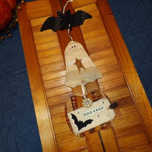May include: A Halloween-themed hanging decoration featuring a black bat with white polka dots, a ghost with a star, and a pumpkin with a "BOO-CREW" tag and a bat silhouette. The decorations are made of fabric and are suspended from a white string.