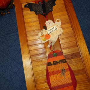 May include: Halloween-themed wooden decoration featuring a bat, ghost, and pumpkin-shaped ornaments. The ghost holds a sign that reads "Tricks & Giggles." The ornaments are made of fabric and have various patterns and colors, including orange, black, and white.